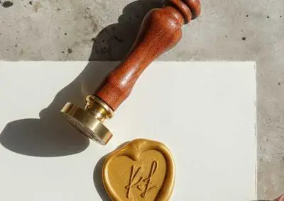 Personalizowany stempel do laku z brązowym trzonkiem, odbicie serca z monogramem KF w kolorze złotym, na białym papierze ślubnym.
