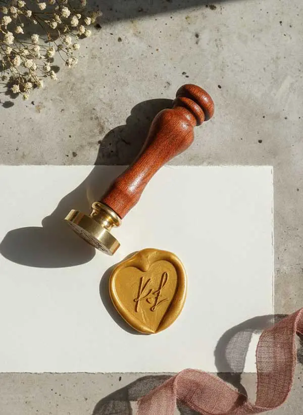 Personalizowany stempel do laku z brązowym trzonkiem, odbicie serca z monogramem KF w kolorze złotym, na białym papierze ślubnym.