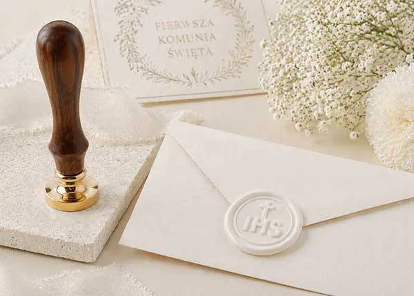 Biała pieczęć do laku z monogramem IHS na eleganckiej kopercie, obok drewnianego gryfu i kwiatów gipsówki