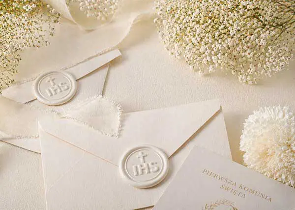 Białe odbicia lakowe z monogramem IHS na kopertach komunijnych w otoczeniu delikatnych kwiatów gipsówki i jedwabnych wstążek.