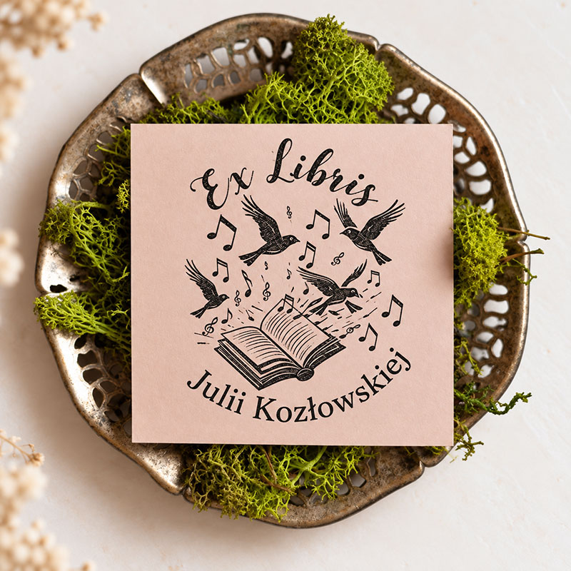Czarne odbicie pieczątki Ex Libris z ptakami, nutami i otwartą książką na pudroworóżowej kartce ułożonej na metalowej tacce z zielonym mchem