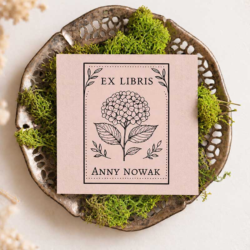Pudroworóżowa kartka z czarnym odbiciem Ex Libris, ilustracją hortensji, napisem Anny Nowak i prostokątną ozdobną ramką, ułożona na metalowej tacce z zielonym mchem