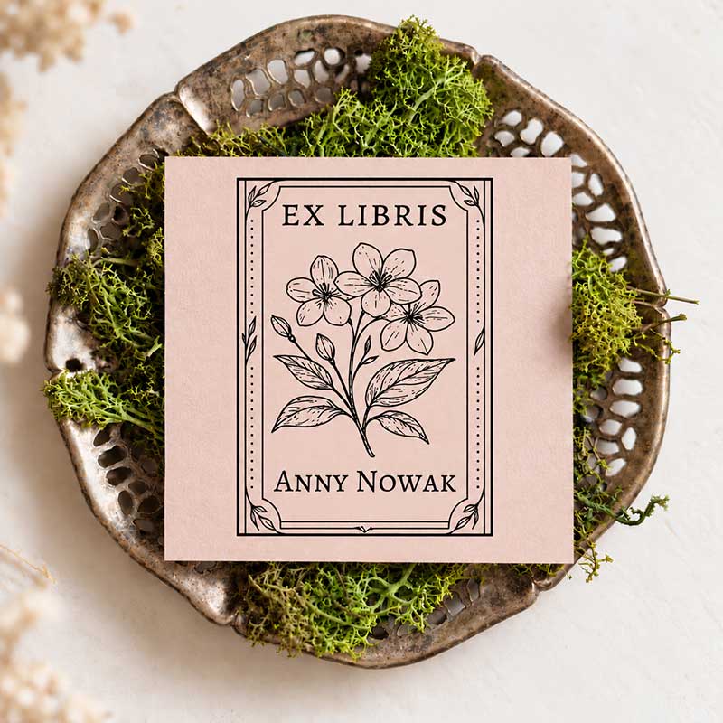 Pudroworóżowa kartka z czarnym odbiciem Ex Libris, ilustracją kwitnącego jaśminu i napisem Anny Nowak w ozdobnej prostokątnej ramce, na metalowej tacce z zielonym mchem
