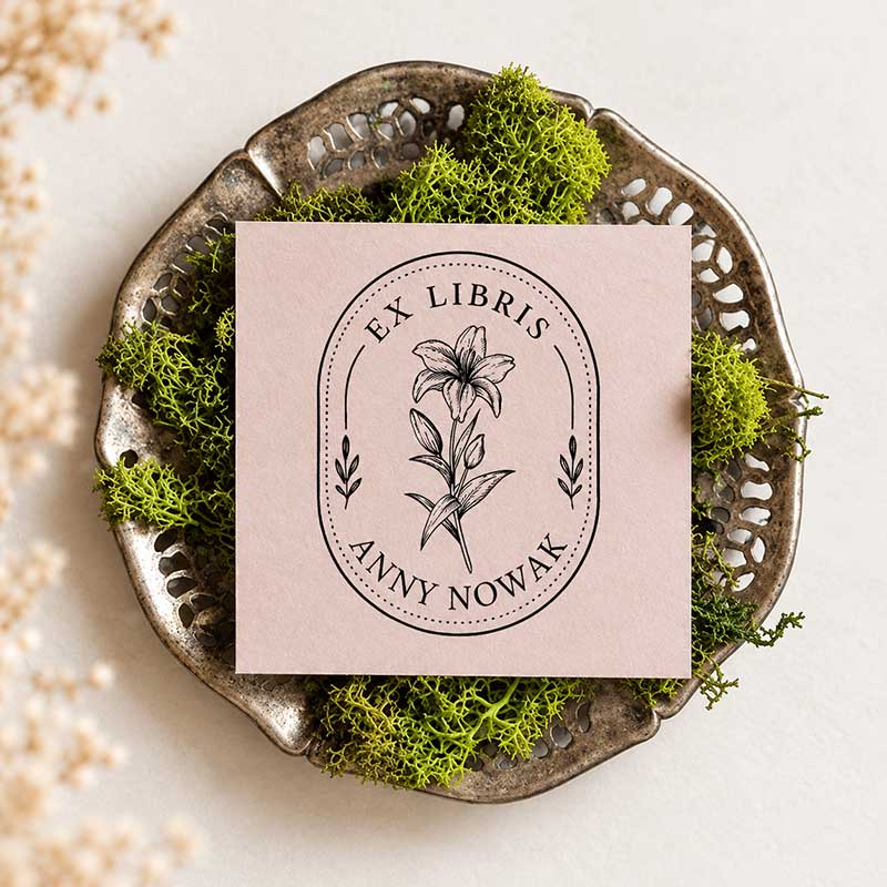 Różowa kartka z czarnym odbiciem pieczątki Ex Libris z kwiatem lilii w owalnej ramce i napisem Anny Nowak, ułożona na postarzanej metalowej tacy z zielonym mchem.